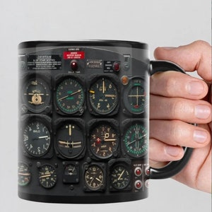 May include: Black ceramic mug with a design of a vintage airplane cockpit instrument panel. The panel features multiple round gauges with black and white markings and red and green needles. The mug has a black handle.