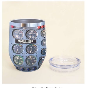 May include: A stainless steel travel mug with a design of an airplane cockpit instrument panel. The panel features gauges, dials, and switches. The mug has a lid and a clear plastic base. The text "NAME HERE U.S. AIRWAYS" is printed on the mug.