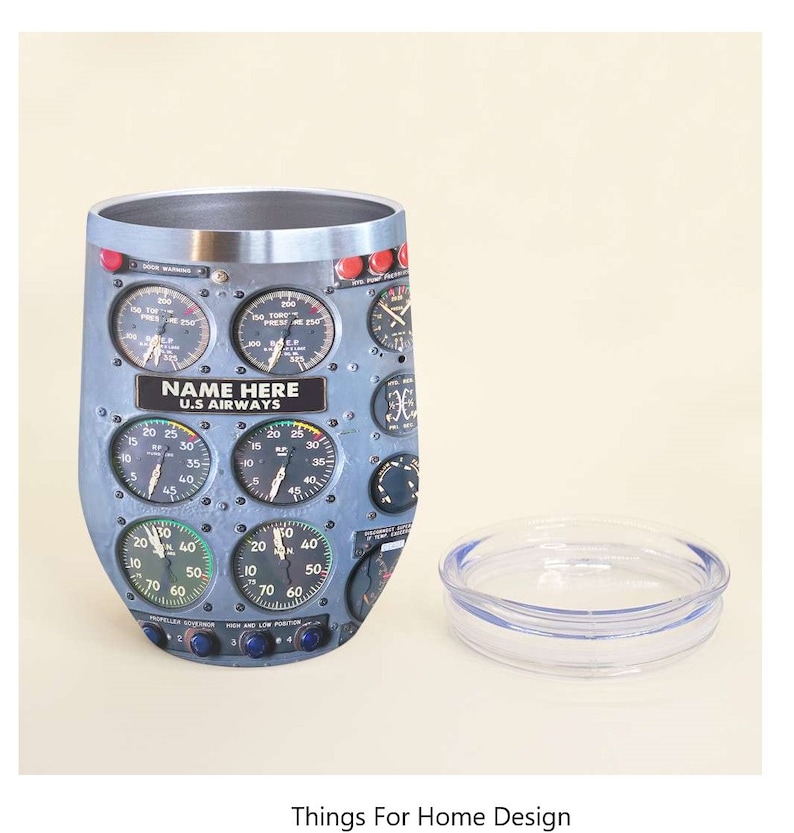 May include: A stainless steel travel mug with a design of an airplane cockpit instrument panel. The panel features gauges, dials, and switches. The mug has a lid and a clear plastic base. The text "NAME HERE U.S. AIRWAYS" is printed on the mug.