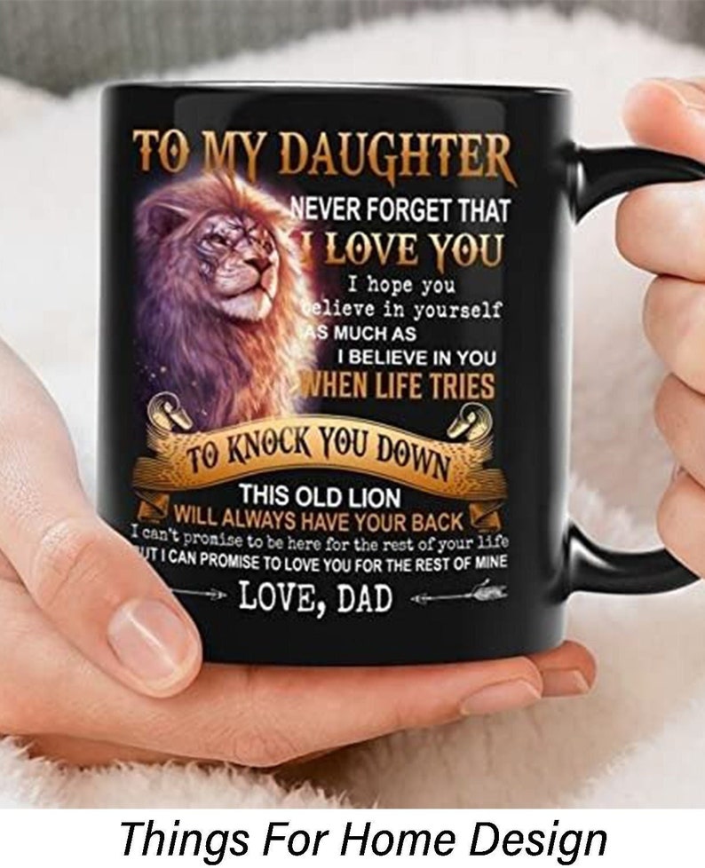 May include: Black ceramic mug with a lion graphic and the text "TO MY DAUGHTER, NEVER FORGET THAT I LOVE YOU." The mug also includes the text "LOVE, DAD." The mug is held by a person.