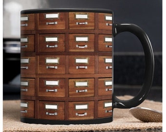 Puede incluir: Taza de café de cerámica negra con un diseño de un armario de madera con cajones. Los cajones tienen tiradores de latón y etiquetas blancas.