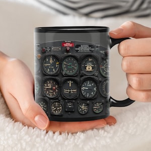 May include: Black coffee mug with a design of an airplane cockpit instrument panel. The panel features various gauges and dials, including a compass, altitude indicator, and airspeed indicator. The text "WARNING CRITICAL MACH No.1" is visible on the panel.