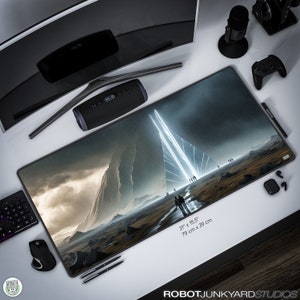 May include: A large, dark grey, rectangular mouse mat with a futuristic landscape scene. The scene features a large, rocky mountain range with a bright white light shining through the centre. The mouse mat measures 31 inches by 15.5 inches, or 79 cm by 39 cm. The text "ROBOT JUNKYARD STUDIOS" is printed in white at the bottom of the mouse mat.