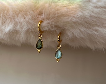 May include: A pair of gold-colored hoop earrings with teardrop-shaped labradorite pendants. The iridescent blue-green stones are accented with small gold beads at the bottom. The earrings are displayed on a fluffy, white surface.