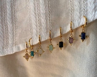 May include: A collection of gold-colored hoop earrings, each featuring a small, square gemstone in various colors, including pink, blue, green, and black. The earrings are displayed against a white, textured fabric.
