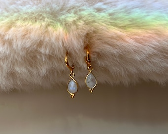 May include: A pair of gold-colored earrings with teardrop-shaped, iridescent stones. The earrings are suspended from small hoops and feature a small gold bead at the bottom of the teardrop. The background is a soft, fluffy, off-white material with a rainbow-colored light.