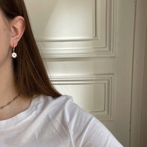 May include: A close-up of a gold hoop earring with a white daisy pendant. The daisy has a yellow center and white petals. The earring is worn on a person's ear. A white t-shirt is visible.