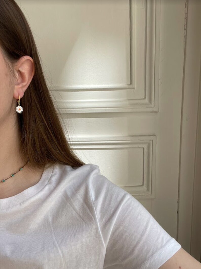 May include: A close-up of a gold hoop earring with a white daisy pendant. The daisy has a yellow center and white petals. The earring is worn on a person's ear. A white t-shirt is visible.