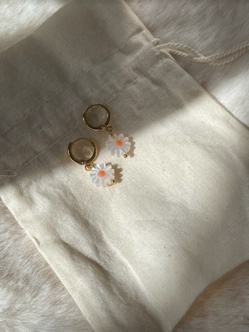 May include: A pair of gold-colored hoop earrings with daisy flower charms. The daisy charms have white petals and a small orange center. The earrings are displayed on a cream-colored fabric.