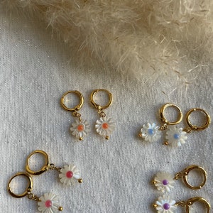May include: Gold-colored hoop earrings with daisy flower pendants. The daisy pendants have white petals and colorful centers in shades of pink, orange, blue, and purple. The earrings are displayed on a textured, off-white surface.