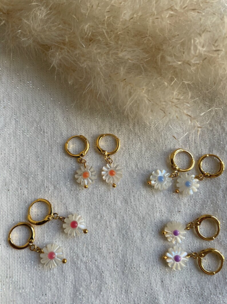 May include: Gold-colored hoop earrings with daisy flower pendants. The daisy pendants have white petals and colorful centers in shades of pink, orange, blue, and purple. The earrings are displayed on a textured, off-white surface.