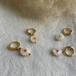 May include: Four gold-colored hoop earrings with white daisy pendants. Each daisy has a different colored center: orange, pink, purple, and blue. The earrings are displayed on a white, glittery surface.