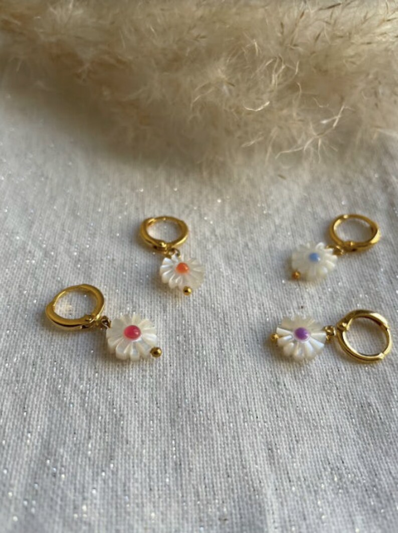 May include: Four gold-colored hoop earrings with white daisy pendants. Each daisy has a different colored center: orange, pink, purple, and blue. The earrings are displayed on a white, glittery surface.