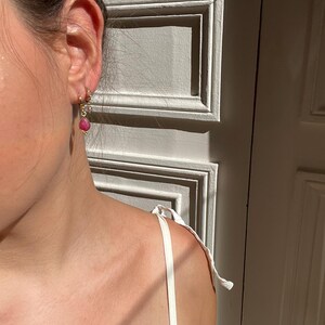 May include: Gold hoop earrings with a dangling pink gemstone and small clear stones. The earrings are worn on an ear, with a white strap of a top visible. The background is a white door with rectangular panels.
