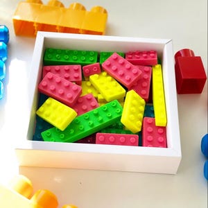 Può includere: Una scatola bianca piena di blocchi di costruzione in plastica colorati. I blocchi sono in varie tonalità di rosso, verde, giallo e blu. La scatola è appoggiata su una superficie bianca.