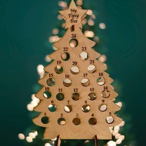 May include: Wooden Christmas tree shaped advent calendar with 24 numbered circles for holding small gifts or treats. The tree is labeled "My Tipsy Tree".
