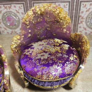 May include: A miniature purple velvet chair with gold accents and a floral patterned cushion. The chair is made of resin and has a unique, ornate design.