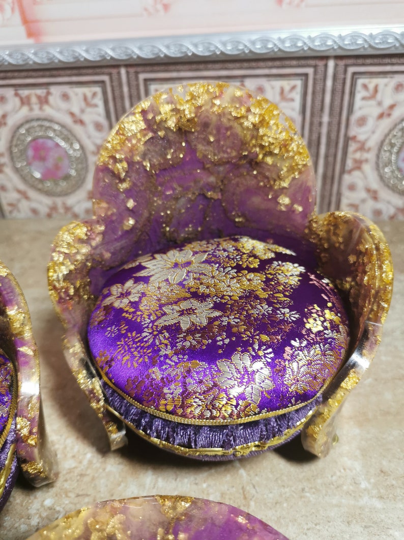May include: A miniature purple velvet chair with gold accents and a floral patterned cushion. The chair is made of resin and has a unique, ornate design.