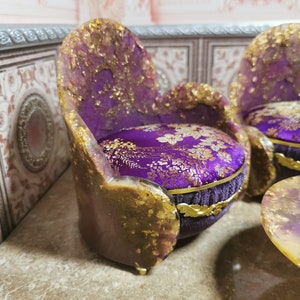 May include: A set of miniature purple and gold chairs with floral patterned cushions. The chairs are made of resin and have a distressed finish.