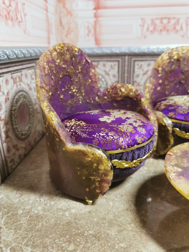 May include: A set of miniature purple and gold chairs with floral patterned cushions. The chairs are made of resin and have a distressed finish.