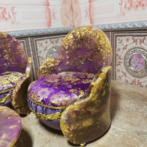 May include: Two miniature purple and gold chairs with a purple and gold patterned fabric cushion. The chairs are sitting on a brown surface.