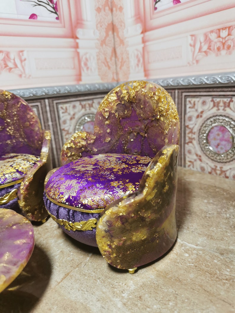 May include: Two miniature purple and gold chairs with a purple and gold patterned fabric cushion. The chairs are sitting on a brown surface.