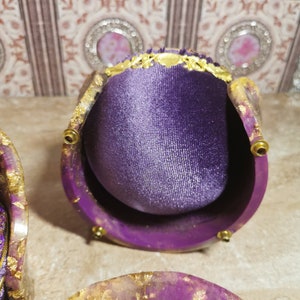 May include: A purple velvet ring box with gold accents and a decorative lid. The box is open and the interior is lined with purple velvet. The box is sitting on a table with a purple and gold resin coaster.