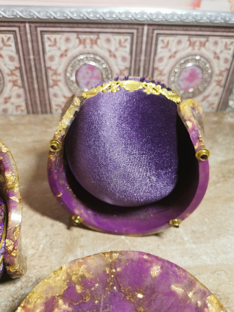 May include: A purple velvet ring box with gold accents and a decorative lid. The box is open and the interior is lined with purple velvet. The box is sitting on a table with a purple and gold resin coaster.