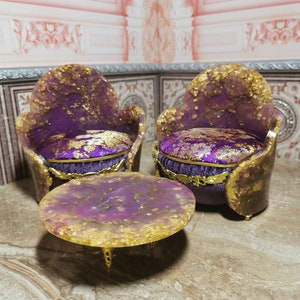 May include: A set of two purple and gold chairs with a matching round table. The chairs have a velvet cushion and the table has a gold base.