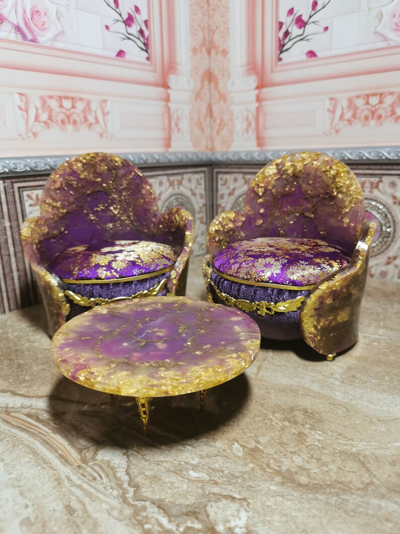 May include: A set of two purple and gold chairs with a matching round table. The chairs have a velvet cushion and the table has a gold base.
