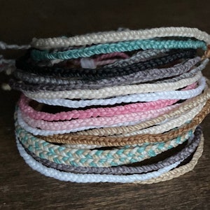May include: A stack of braided bracelets in various colors, including white, pink, gray, and teal. The bracelets are made of a woven material and have adjustable closures. The stack is on a dark wooden surface.