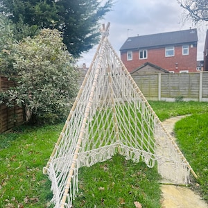 Op de afbeelding: Een witte macramé tipi met houten palen, opgesteld in een grasachtige achtertuin.