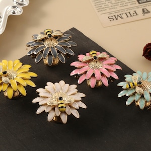May include: A selection of flower-shaped cabinet knobs in shades of yellow, pink, and blue. Each knob features a bee design. The knobs are made of metal and are likely used for furniture, with measurements in centimetres.