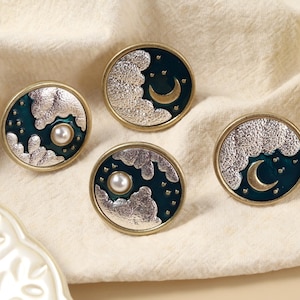 May include: Four round cabinet knobs with a celestial design. Each knob features a silver cloud, a dark teal background, and a gold crescent moon or pearl. The knobs have a gold-coloured rim.