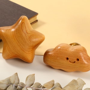 Puede incluir: Pomos de armario de madera con forma de estrella y nube. La estrella tiene cinco puntas y la nube tiene una cara sonriente. Ambos son de color marrón claro con vetas de madera. También se ve un globo terráqueo de madera sobre una base de latón.