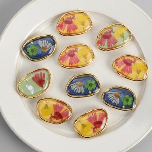 May include: Ten oval brooches with gold edging, each featuring a pressed flower design in resin. The flowers are in pink, yellow, blue, and green. The brooches are displayed on a white plate. The brooches are approximately 2.5 cm in length.