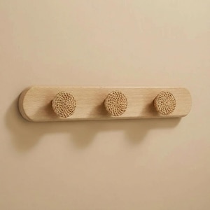 May include: A wooden wall-mounted coat rack with three round, woven rattan hooks. The rack is made of light-colored wood and is mounted on a neutral-colored wall. The hooks are evenly spaced along the wooden board.