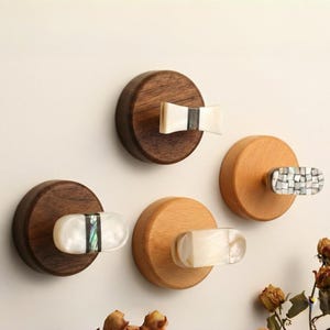 Op de afbeelding: Een verzameling decoratieve wandhaken. De haken hebben ronde houten bases in verschillende tinten bruin en licht hout. Elke haak heeft een uniek decoratief element, waaronder een vlinderdasvorm, mozaïek en ovale ontwerpen, allemaal in wit en iriserende materialen.