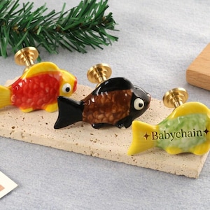 Könnte beinhalten: Drei bunte Keramik-Schrankknöpfe in Fischform mit Messingbeschlägen. Die Fische sind rot, braun und gelb mit schwarzen Akzenten. Der Text "Babychain" befindet sich auf einem der Fische. Sie werden auf einem rechteckigen Stein ausgestellt.