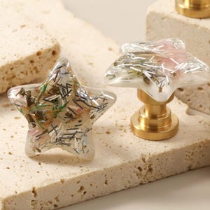 以下が含まれることがあります： 金色のベースが付いた星型のキャビネットノブ。ノブには、銀と緑のグリッターと小さなピンクの花が詰められています。ノブは、明るい色のテクスチャのある表面に表示されています。