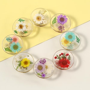 May include: A collection of eight clear, round resin coasters, each featuring a different pressed flower arrangement. The flowers include daisies, purple, red, yellow, and blue blooms, with green foliage. The coasters are arranged in a circular pattern.