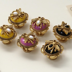 Peut inclure: Boutons de meuble décoratifs avec un motif d'abeille. Les boutons ont une base en métal doré et un centre coloré en jaune, violet et noir. Chaque bouton présente un accent d'abeille et une pierre centrale blanche ou noire.