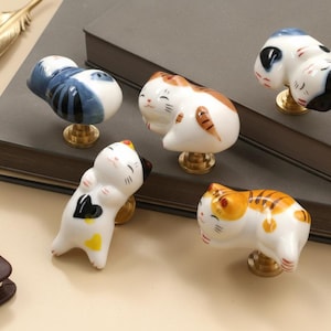 Könnte beinhalten: Eine Sammlung von keramischen Katzen-Schrankknöpfen in verschiedenen Posen und Farben, darunter Weiß mit blauen, orangefarbenen und schwarzen Akzenten. Jeder Knopf hat einen goldfarbenen Sockel, für Möbel.