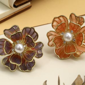 May include: Two floral-shaped brooches. One is a deep purple and gold, the other is orange and gold. Each has a pearl center and small crystal accents. The brooches are set against a neutral background.