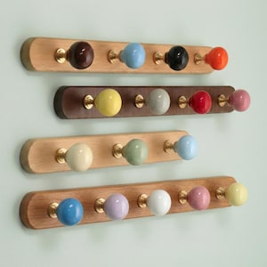 Könnte beinhalten: Vier hölzerne Garderobenleisten mit bunten Keramikknöpfen. Die Leisten sind aus hellem Holz gefertigt und haben messingfarbene Beschläge. Die Knöpfe sind in verschiedenen Farben erhältlich, darunter Braun, Blau, Schwarz, Orange, Gelb, Grau, Rot, Rosa, Grün und Weiß.