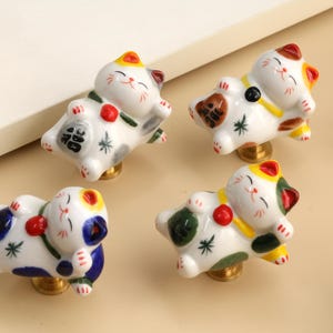 Puede incluir: Un juego de cuatro pomos de cerámica para armarios con forma de gatos Maneki-neko reclinados. Cada gato blanco presenta detalles pintados en negro, naranja, azul y verde, con un collar rojo y una base de metal dorado para fijar a muebles.