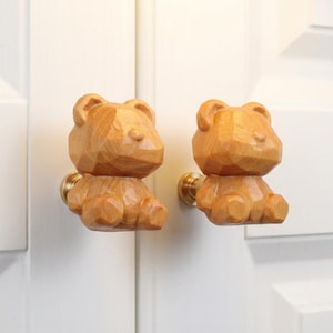 May include: Two wooden bear-shaped cabinet knobs. The knobs are light brown with a geometric design and brass hardware. They are mounted on a white cabinet door. The knobs are a charming addition to any nursery or children's room.