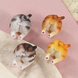 May include: Four cat butt figurines in various colors: gray and white, orange and white, brown and white, and yellow and orange. Each figurine has a tail and pink paw pads. The text "Babychain+" is visible on one of the figurines.