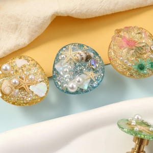 May include: Three decorative resin knobs with beach-themed designs. One is gold with seashells, pearls, and a starfish. Another is blue with starfish, shells, and pearls. The third is gold with pink and green floral accents and a gem.