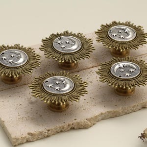 May include: Five decorative cabinet knobs with a celestial design. Each knob features a silver-colored moon and star design set within a sunburst frame. The knobs are made of brass and are displayed on a light-colored stone.